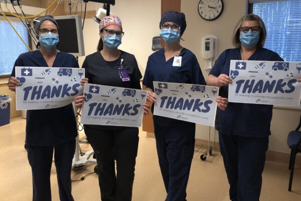 Four women in blue scrubs and masks hold up signs that say Thanks