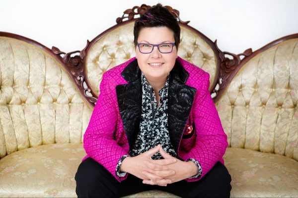A woman in a bright pink jacket and wearing glasses sitting on an antique gold tufted couch.