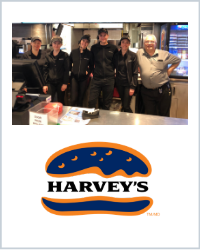 Seven Harveys restaurant employees pose in their black uniforms under the menu boards. The Harvey's Logo is below