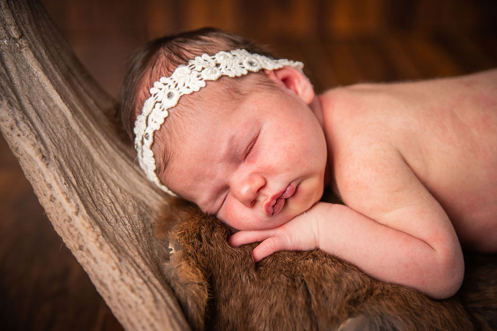 A baby girl in a lace headband sleeping on a brown fuzzy fur.
