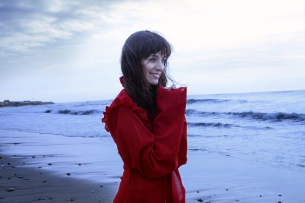 A smiling, dark haired woman in a red coat standing on the beach.