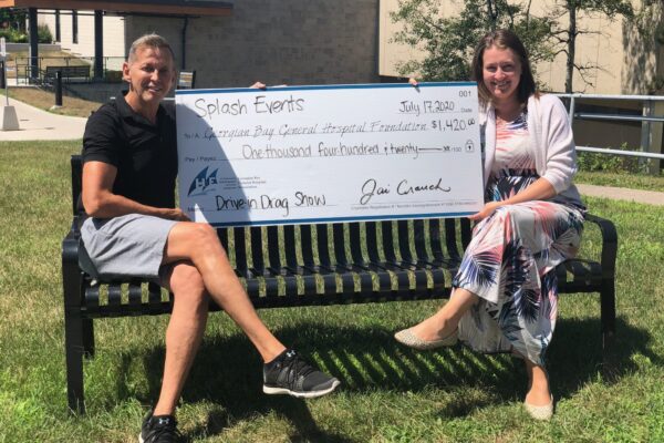 Two women sit on a metal bench holding a giant cheque for $1,420 from the Driven Drag Show to the GBGH Foundation