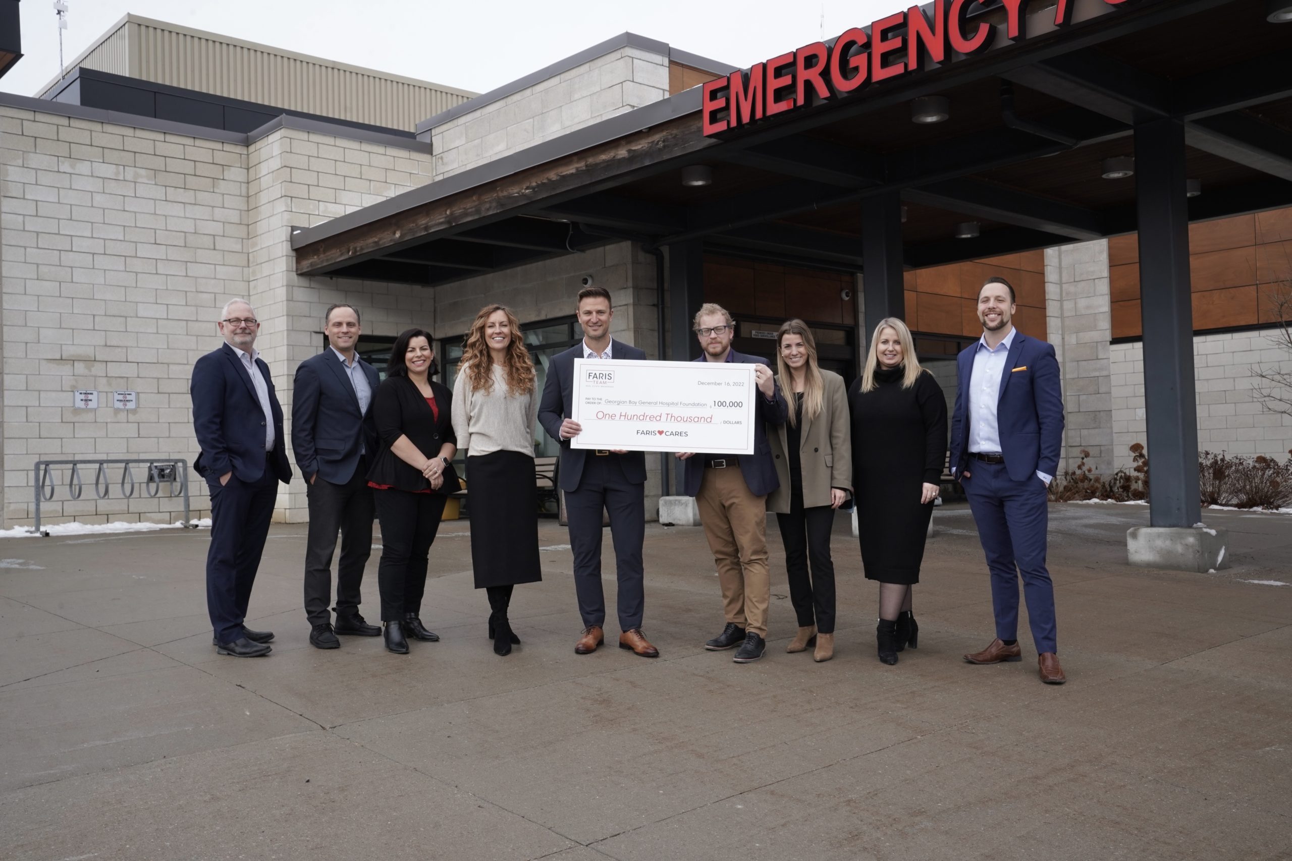 Nine people stand outside the emergency entrance to GBGH holding a giant cheque for $100,000 made out to the GBGH Foundation from the Faris Team.