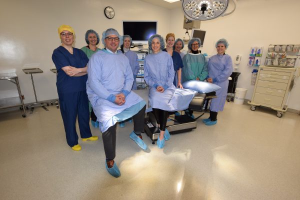 Nine people in either gowns or scrubs gathering around or sitting on a bed in the operating room