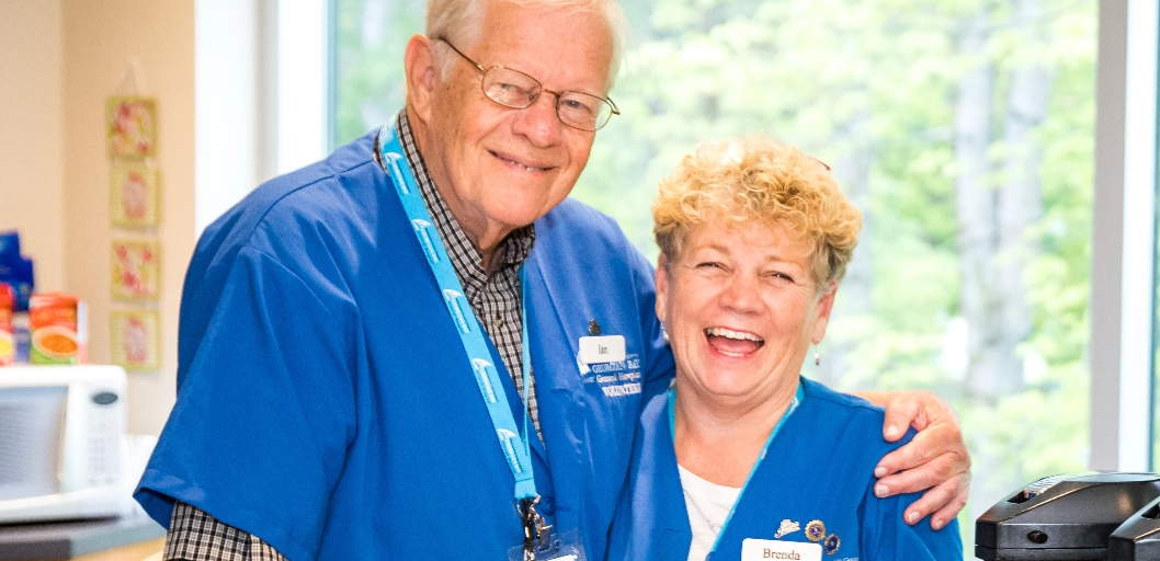 Smiling older male and female in their blue hospital volunteer scrubs.