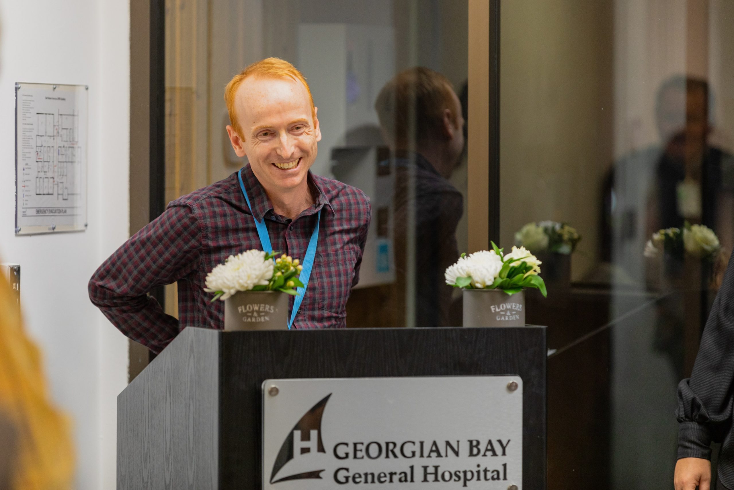 A man with receding red hair wearing a plaid shirt stands at a podium that has Georgian Bay General Hospital written on it