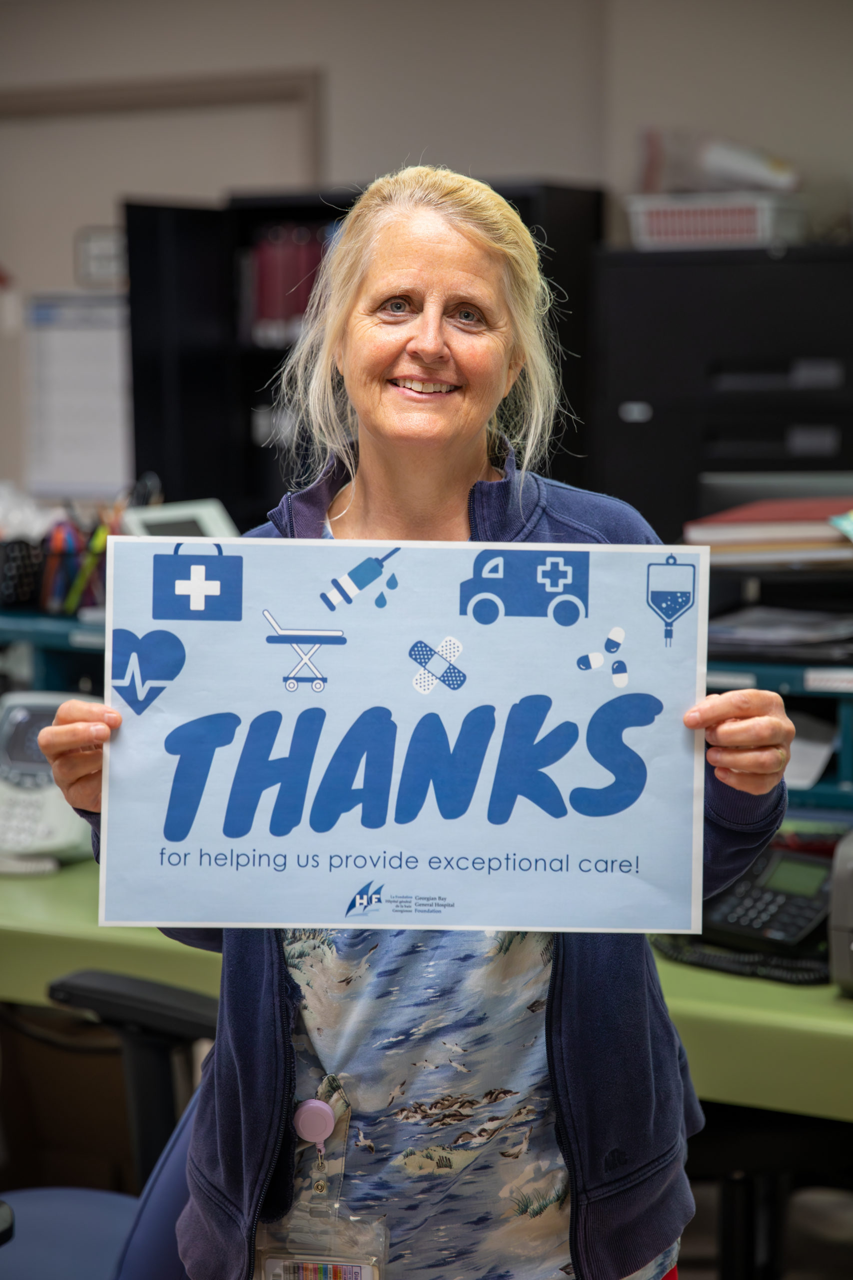 An older woman with blond hair tied back. She holds a sign that says Thanks for helping us provide exceptional health care.