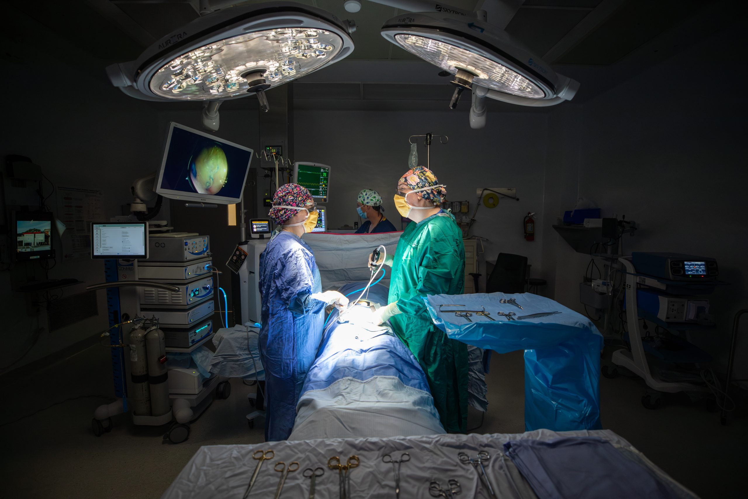 A dark operating room with two people dressed in scrubs, holding surgical instruments.