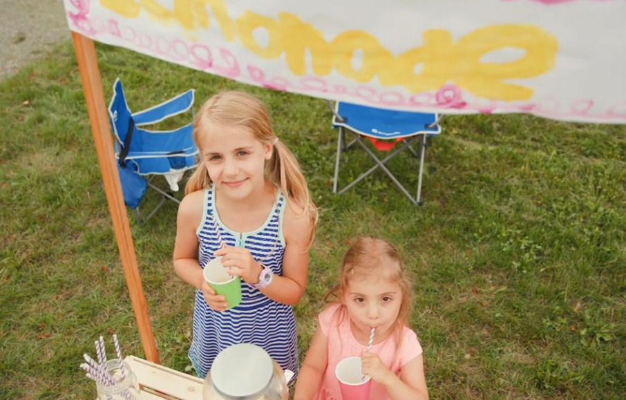 Two girls drinking lemonade with a hand painted sign reading Lemonade above them.