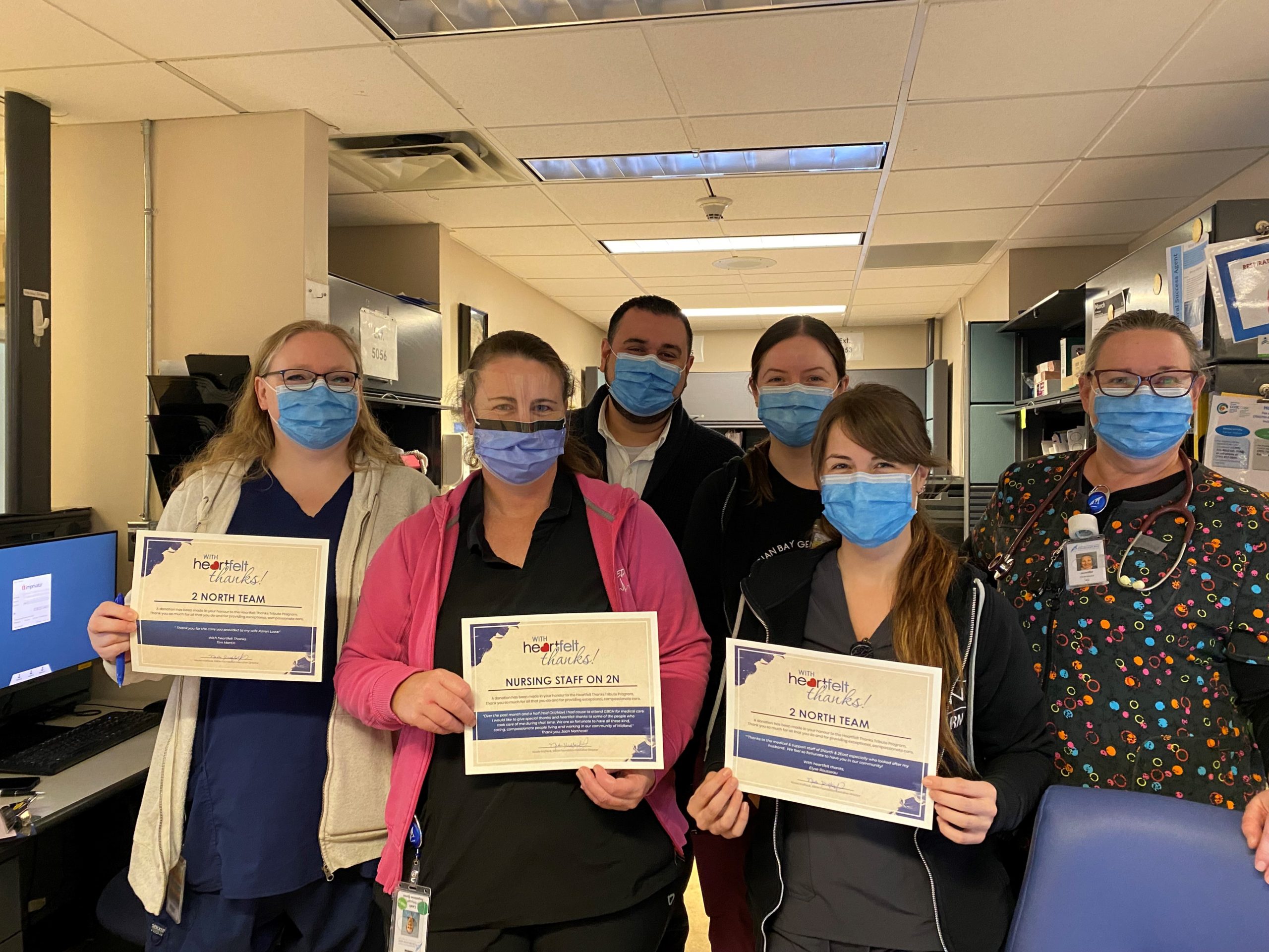 Five woman ans a man all wearing masks stand in one of the health stations. Three of the people hold up Heartfelt Thanks Certificates for the 2 North team members.