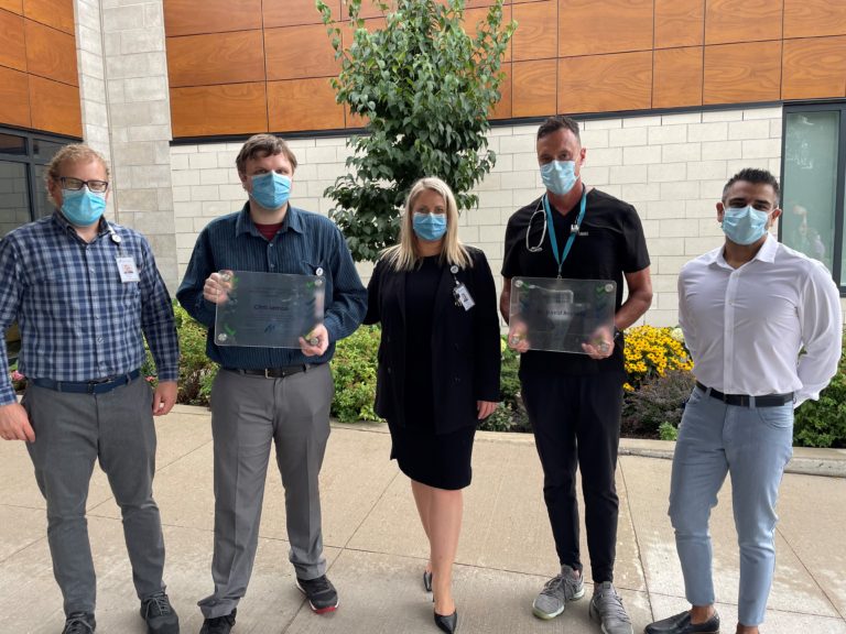 Five people are outside with a garden behind them. Two men are holding clear glass certificates.