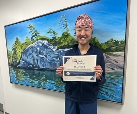 Dr. Amy Huang holding a Hearthfelt Thanks certificate in front of a nice painting of a rock cliff, white pines, and Georgian Bay