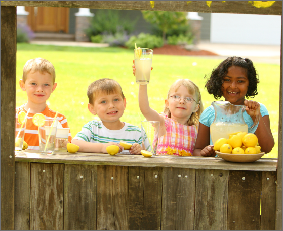 2 young boys and 2 girls standing behind a wooden structure with lemons and a glass jug of lemonade