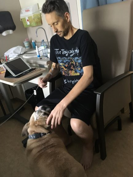 A man in a black band shirt is sitting in a chair in the hospital petting a dog