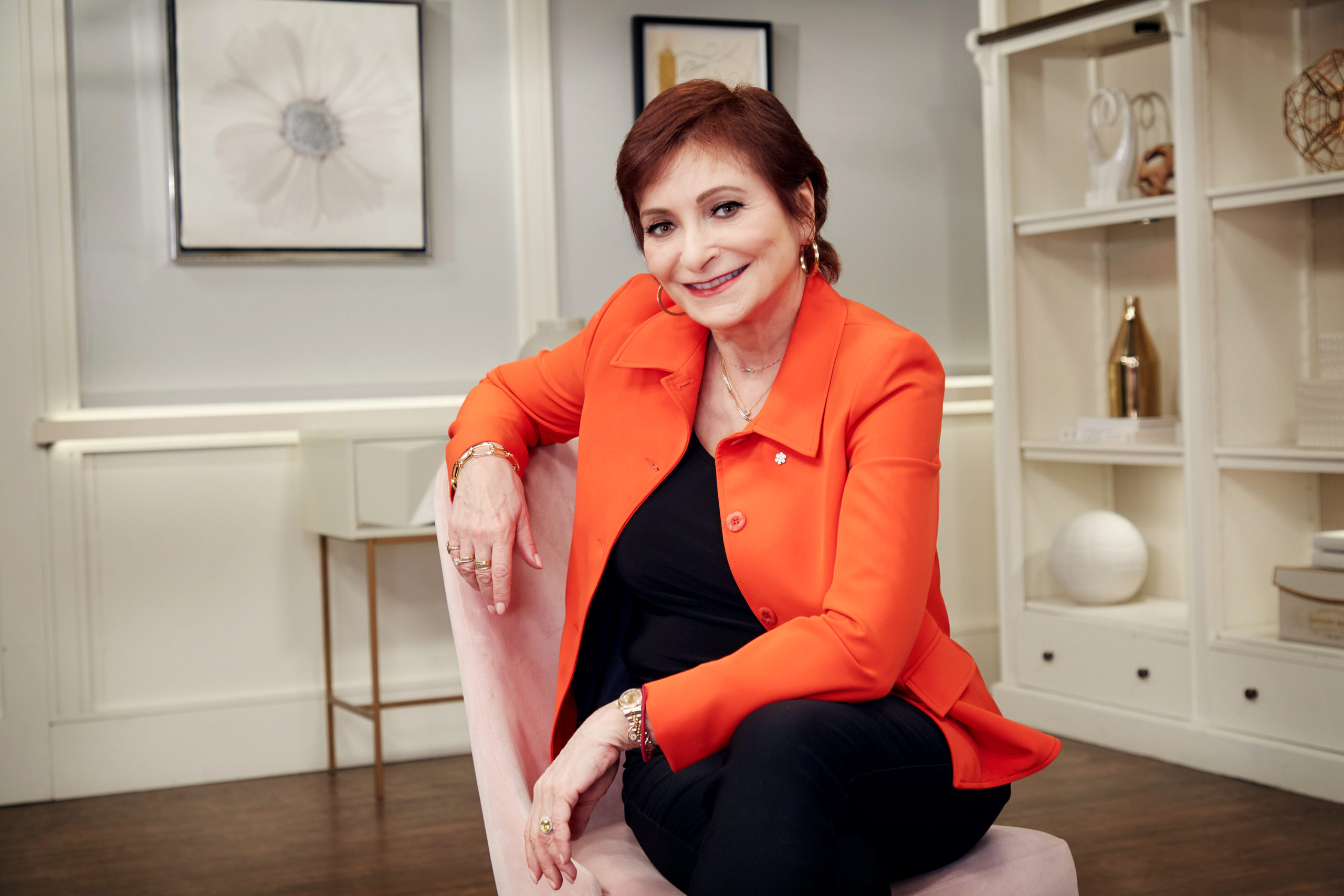 A woman sitting in a pink chair. She has short red hair and is wearing black pants and top with a bright orange suit jacket.