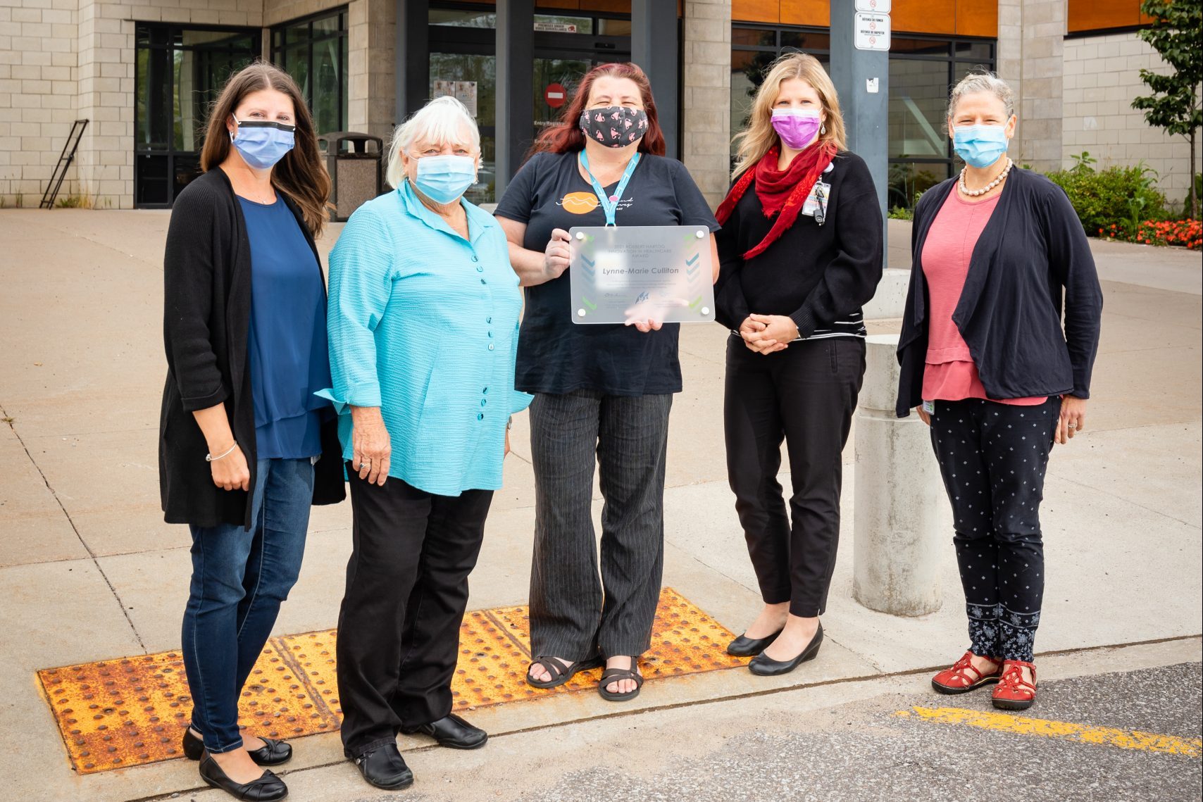 Five women in masks stand outside the hospital. One of them in holding an award plaque