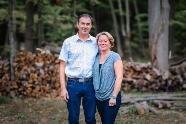 man in a light shirt and jeans and woman in blue top and pants pose in front if a pile of cut logs