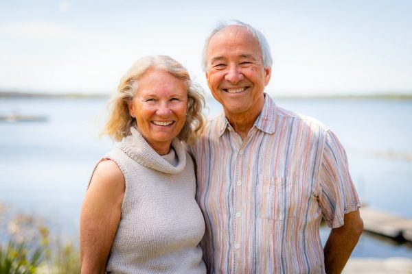 Smiling couple in front of the bay. Blonde women in a grey sleeveless sweater and a man in a blue and orange striped shirt