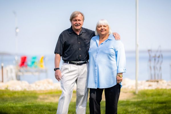 A man in a black shirt and white pants with his arm around a grey haired woman in a blue shirt and black pants. The Bay is behind them