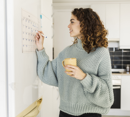 A woman in a green sweater holding a yellow cup writing on a wall calendar