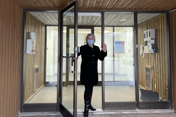 A woman in a mask standing at glass doors waving