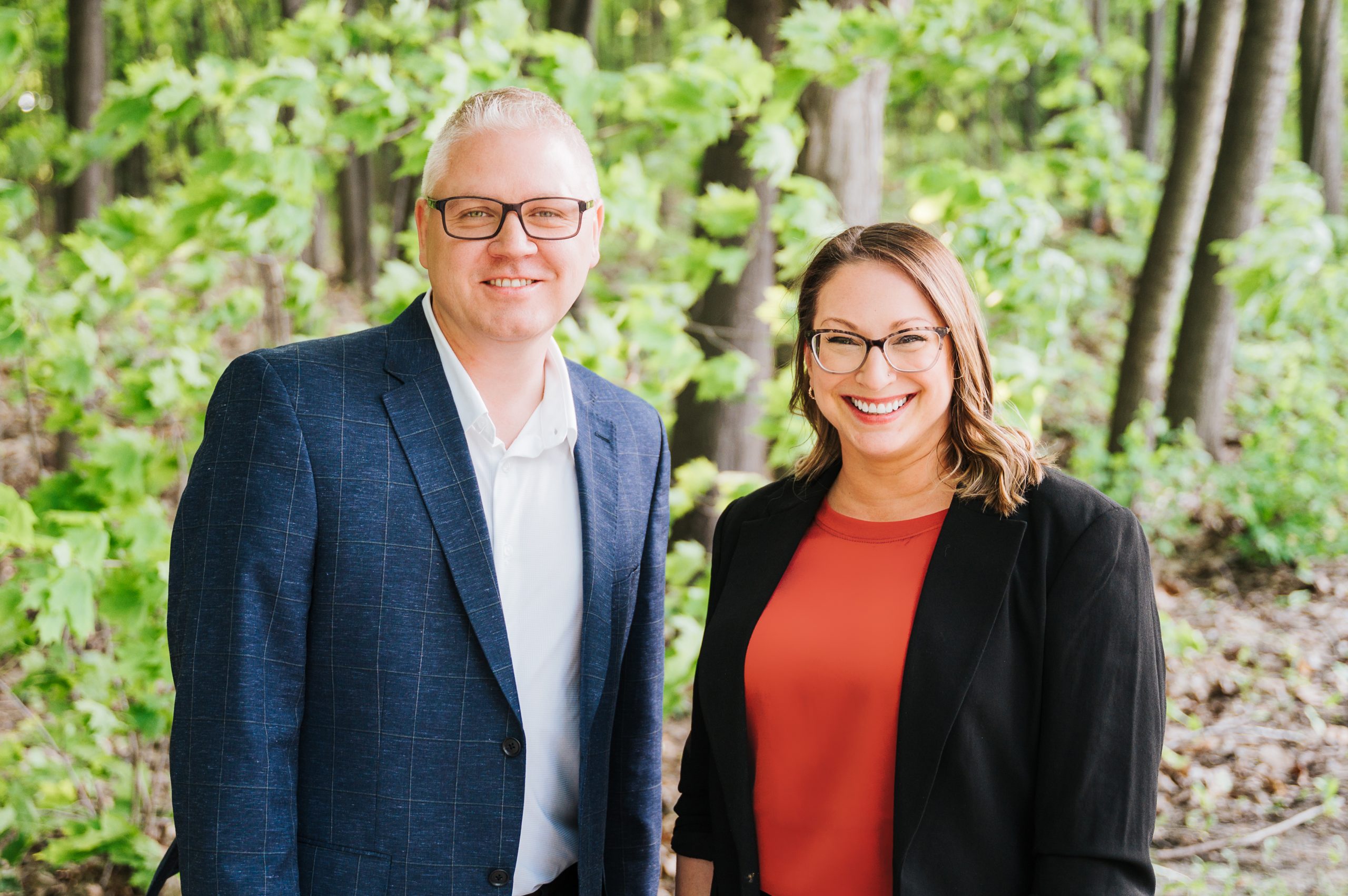 A man and woman pose outdoors with lots of green trees bedhind them