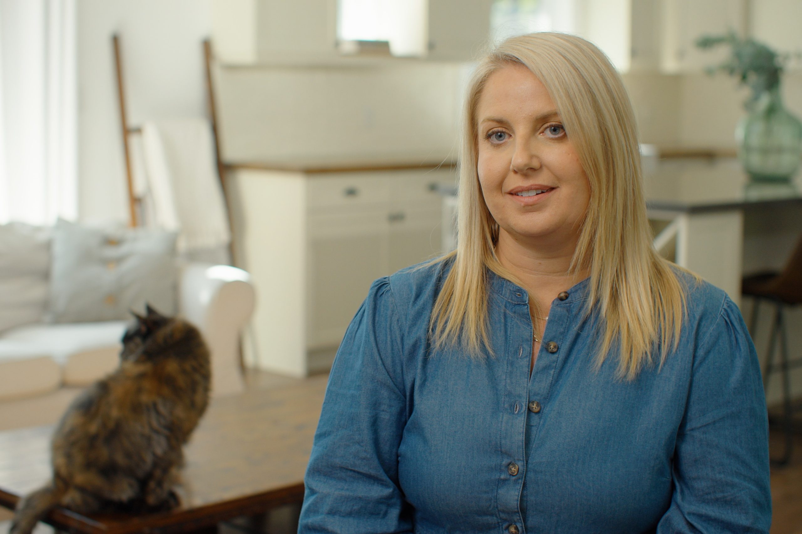 A blond women in a jean coloured top. A brown and red cat sits on the coffee table beside her.
