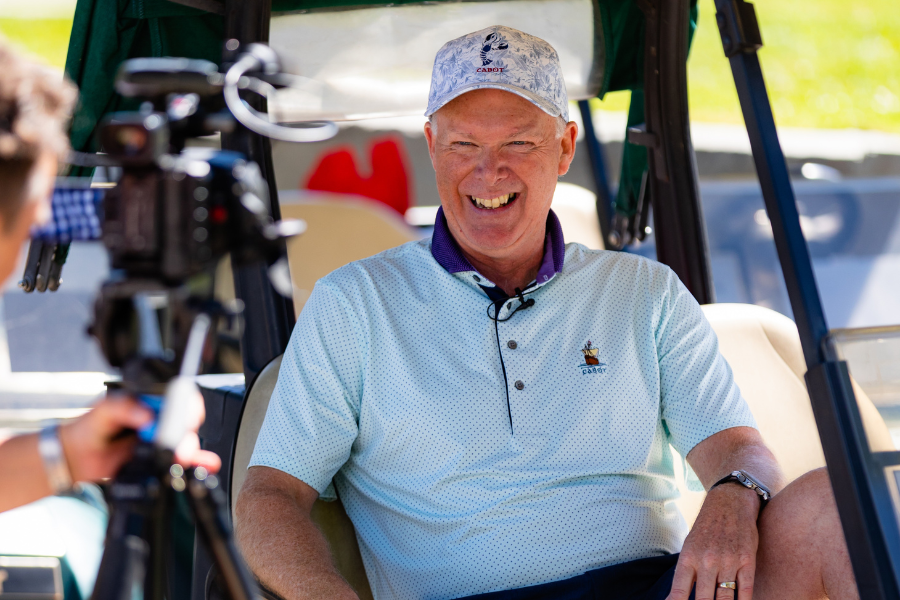 Glenn Howard wearing a blue golf shirt and hat sitting in a golf cart laughing
