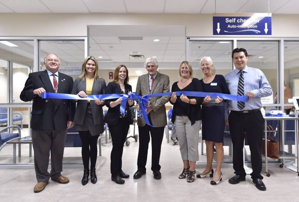 Three men and four woman are all holding up a long blue ribbon with a bow tied in the middle. Behind them is the entrance to the emergency room self-check-in area