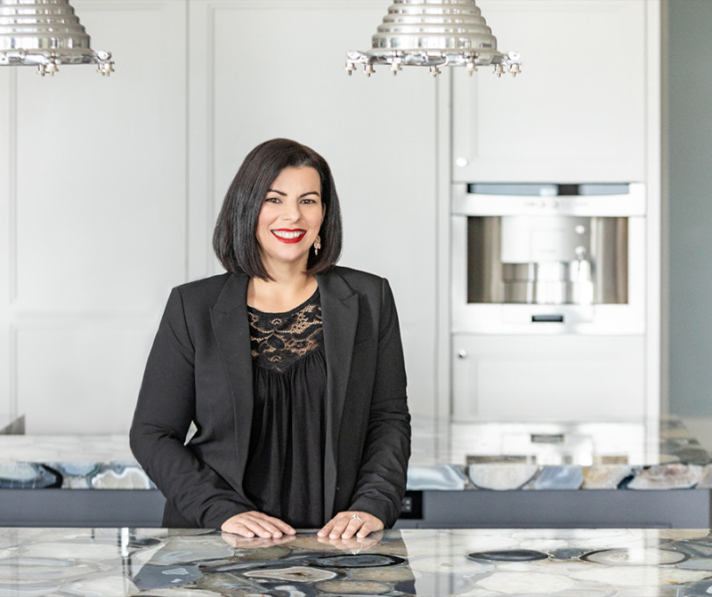 A smiling woman with black hair wearing a black top and black jacket is standing in a high end white kitchen at a marble countertop.