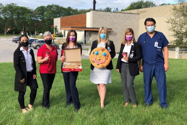 Six people in masks stand outside on the grass. Two hold a Tim Horton's coffee, one a box of cookies and one a giant smile cookie.