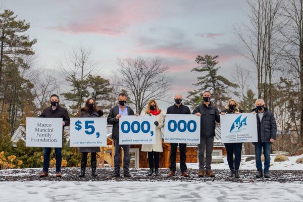Eight people stand outside in winter wearing masks. They hold posters that together say Marco Mancini Family Foundation $5,000