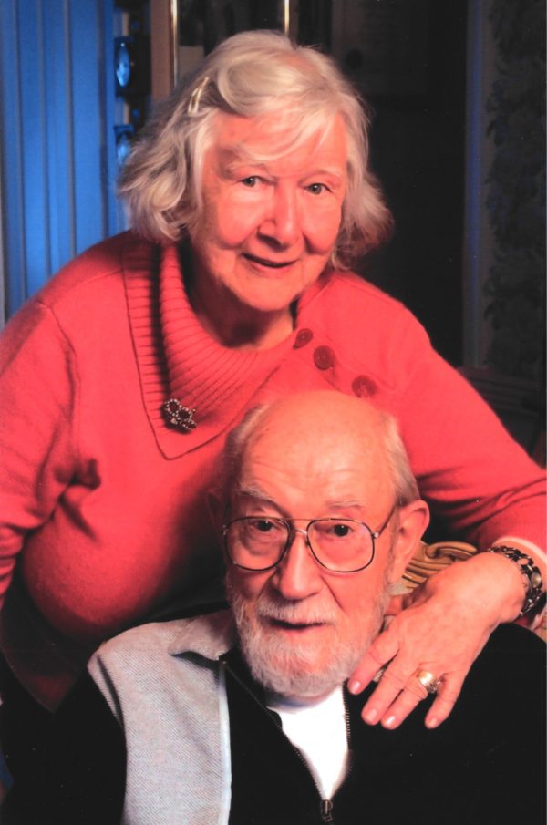 Grey haired woman in a red sweater has a hand on the shoulder of a balding grey haired man with a beard