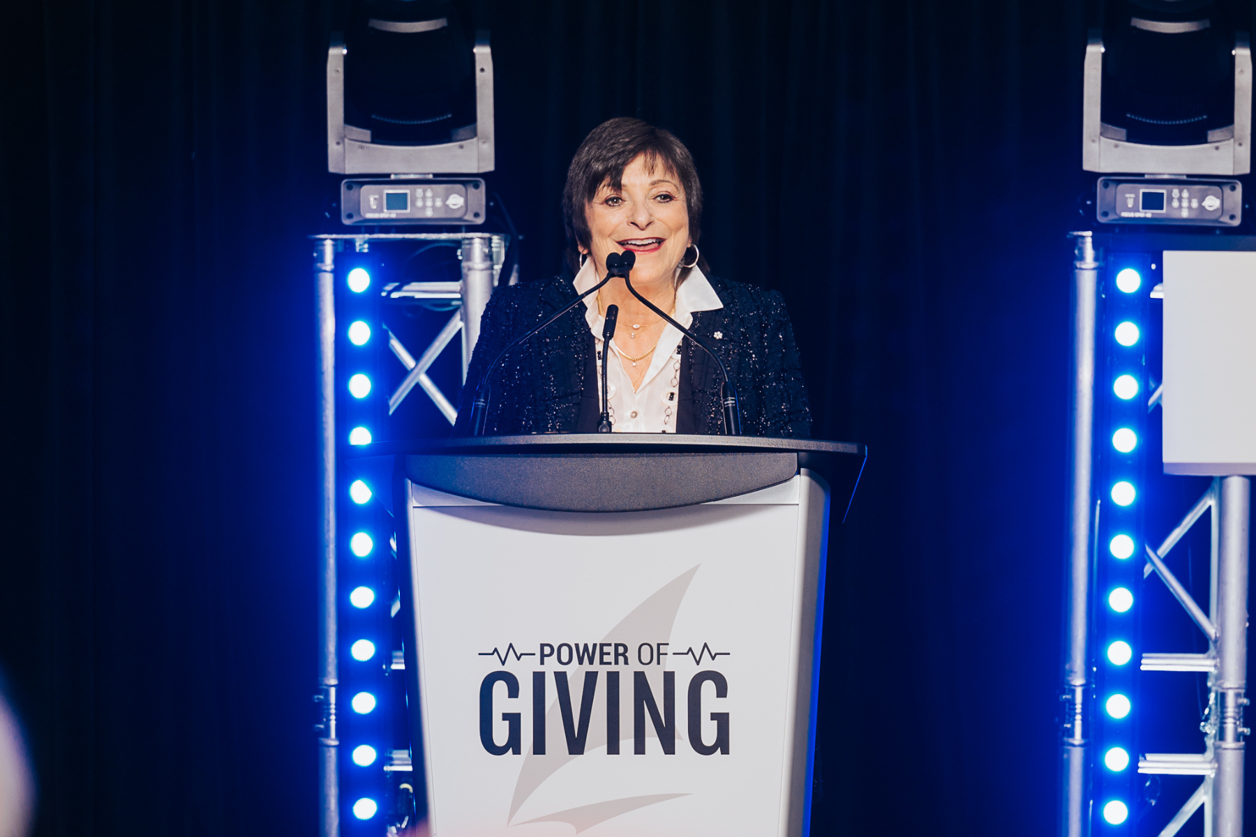 A woman with short dark hair wearing a white shirt and black sparkly jacket is speaking at a podium with the words Power of Giving on it.