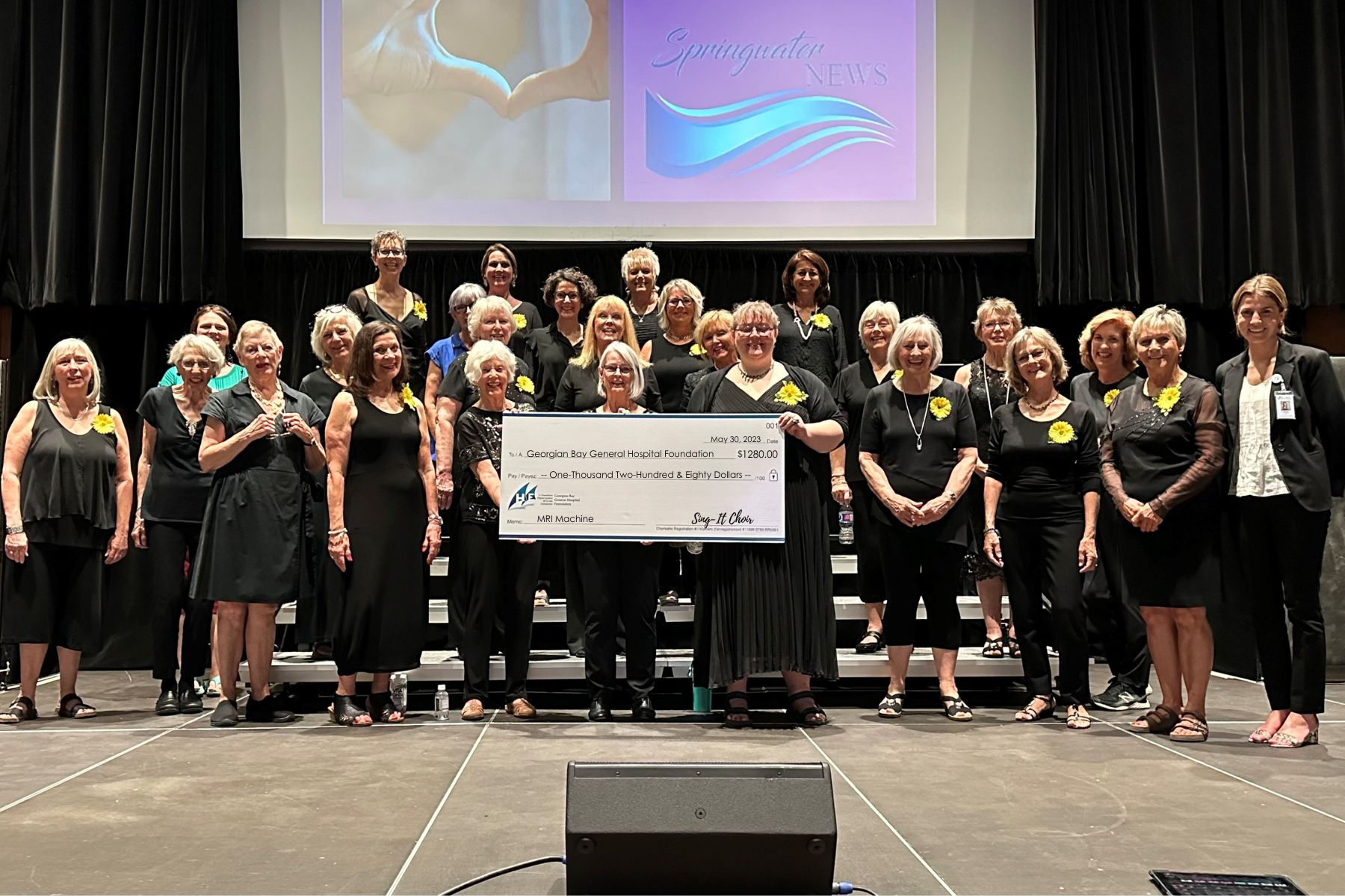 Twenty-four women in black clothing hold a cheque made out to the GBGH Foundation MRI Machine Fund for $1,280.00 from Sing-It choir.