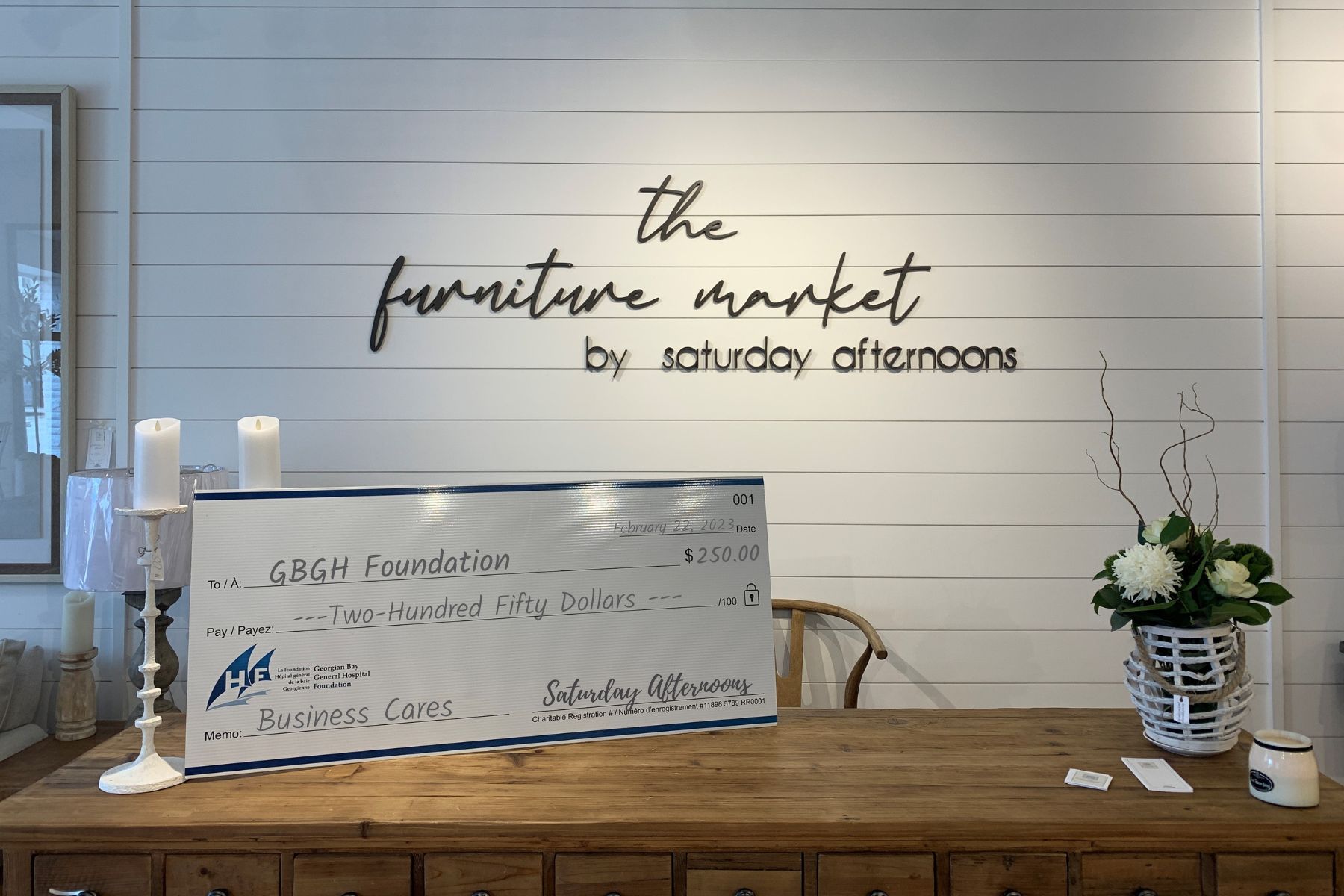 A giant cheque sits on a wooden table. The white wall behind has the words The furniture market by saturday afternoons. The cheque is made out to GBGH Business Cares for $250 from Saturday Afternoons