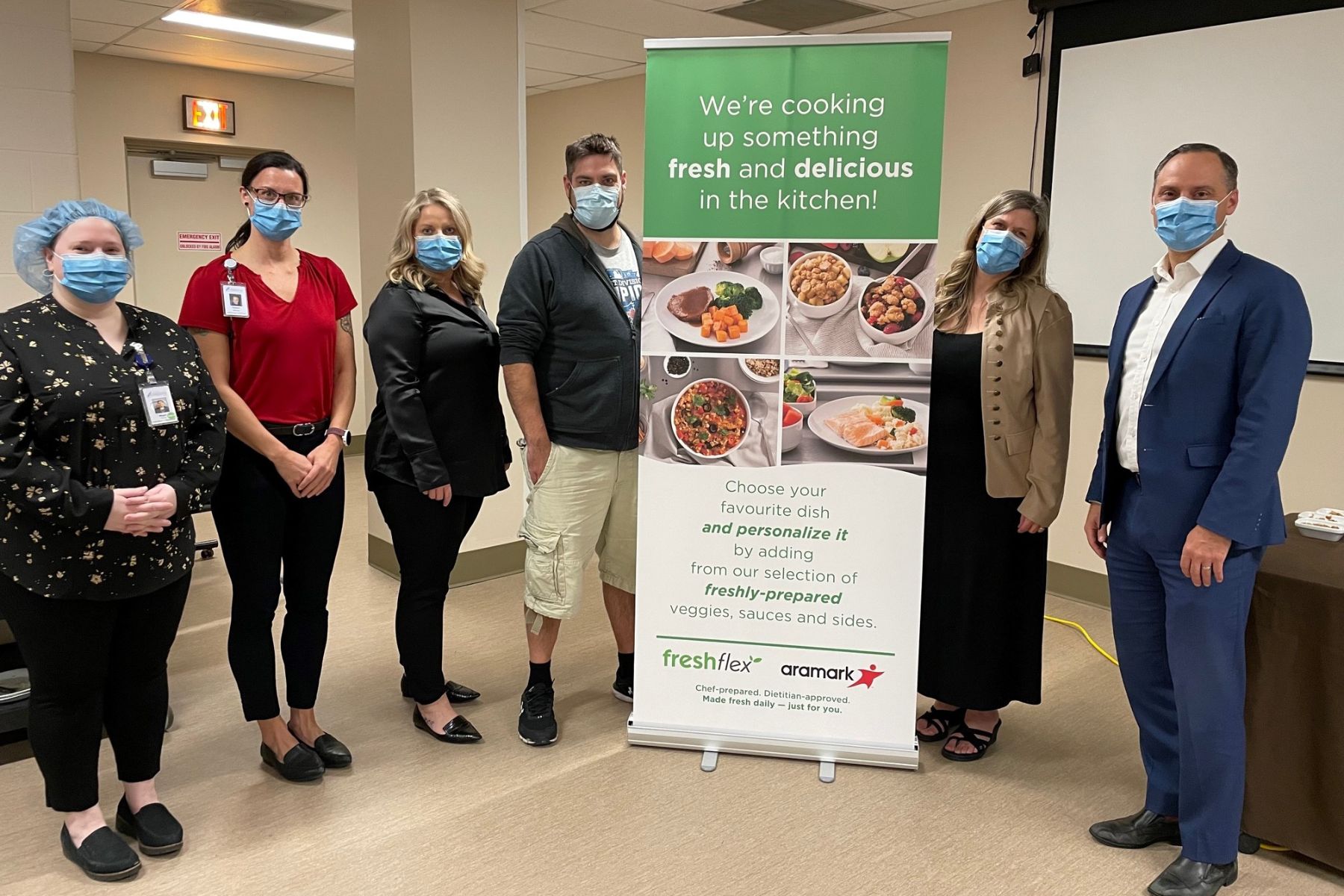 Six hospital workers gather around a a sign with four images of plated food. The sign reads We're Cooking Up something fresh and delicious in the kitchen